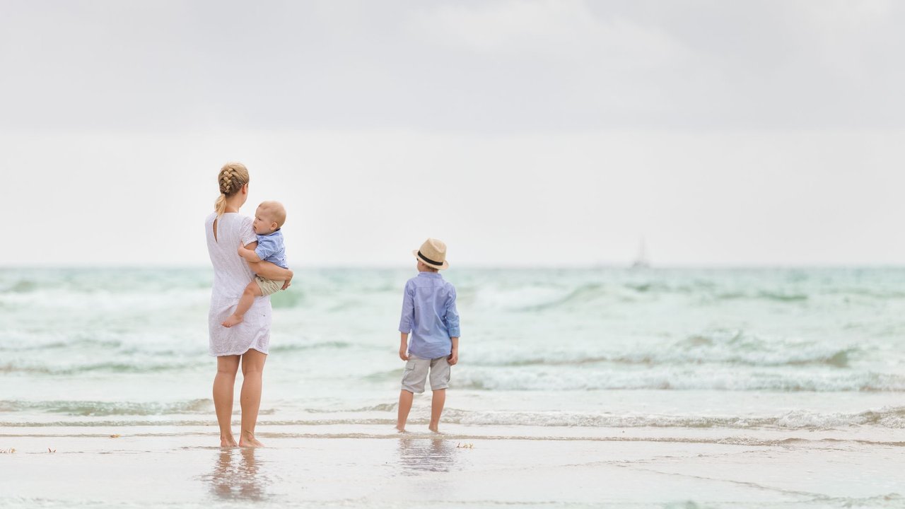 Young mother in white dress walking with her two little boys along the ocean beach. 