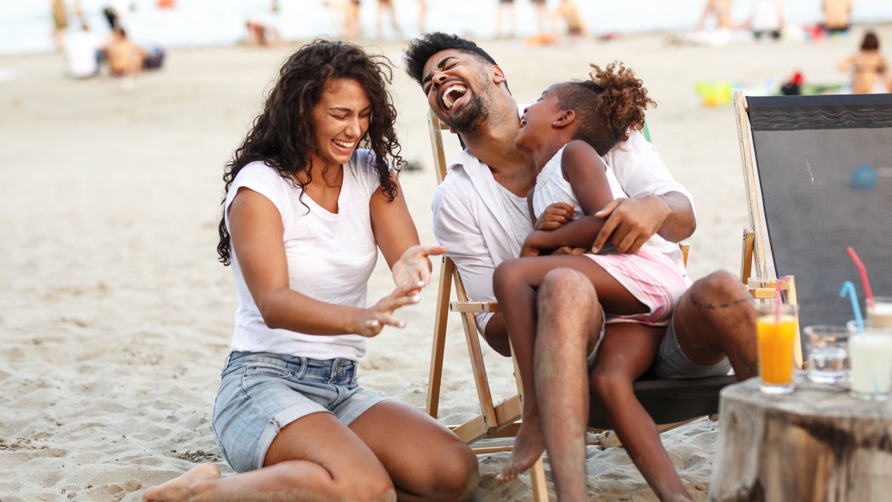 Young mixed race family sitting and relaxing at the beach on beautiful summer day.Daughter lies in father lap and laughing.