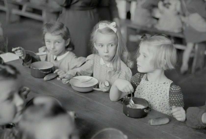 Kinder sitzen in einer schwarz-weiß Aufnahme an einem Tisch und essen. 