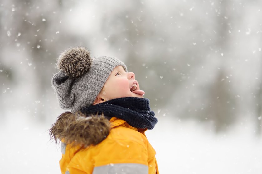 Kinder Weihnachtslieder: Junge in gelber Winterjacke und grauer Mütze fängt Schneeflocken mit dem Mund