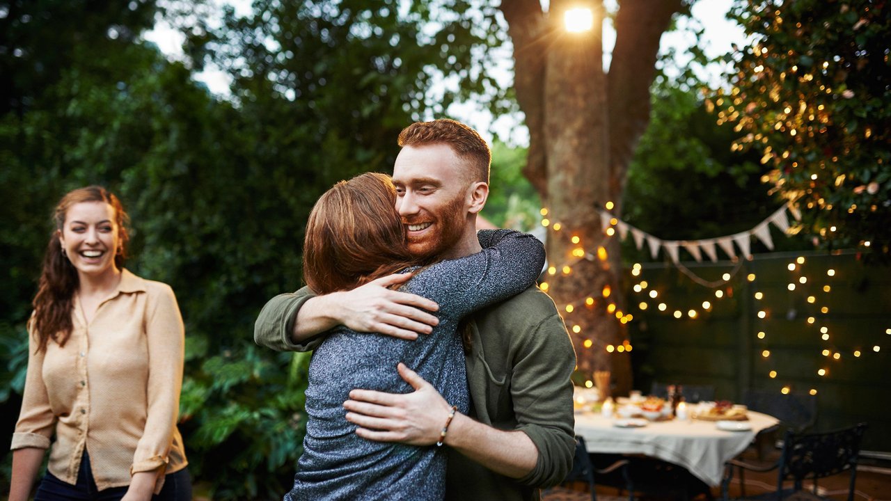 Polterabend-Ablauf: Mann umarmt Frau, andere Frau steht lachend daneben, Garten-Party-Hintergrund