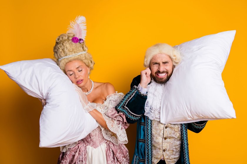 Eine humorvolle Darstellung eines jungen Paares in eleganten Kostümen im königlichen Stil, das weiße Kissen hält. 