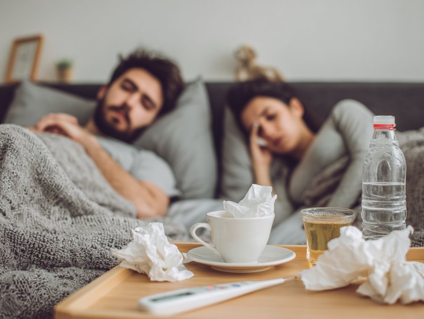 Gemeinsam erkältet: Ein Tisch mit Taschentüchern und Trinken, im Hintergrund ein krankes Paar