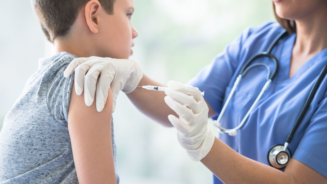 A young Caucasian boy sits up on an exam table with his sleeve over his shoulder, as a female Latin nurse gives him his immunization needle.  The boy is dressed casually in a t-shirt and the nurse is wearing blue scrubs and white medical gloves for protection, as she holds the young boys shoulder with one hand and gives the injection with the other.