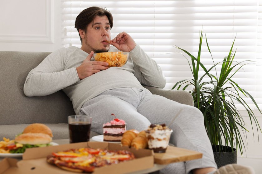 Junger, übergewichtiger Mann isst Fast Food auf dem Sofa