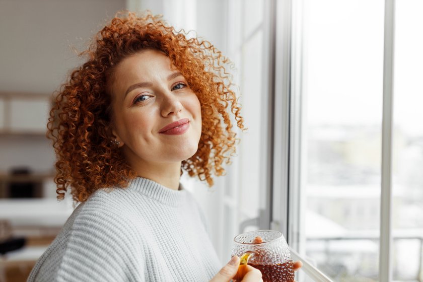 Closeup image of cute attractive millennial redhead woman with funny curls enjoying tea standing next to big panoramic window with city landscape, looking at camera with happy satisfied smile