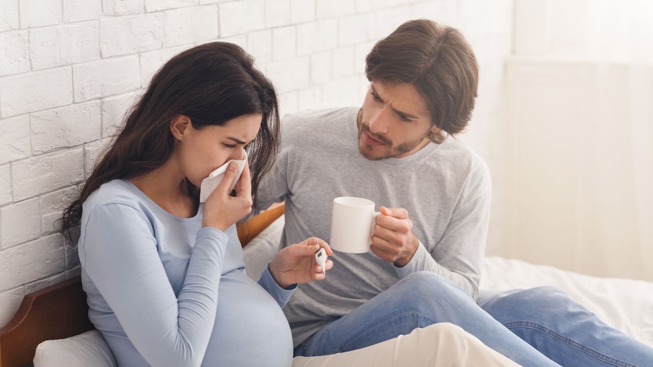 Pregnancy And Cold. Caring husband looking after his sick pregnant wife that suffering from flu, sitting on bed at home together