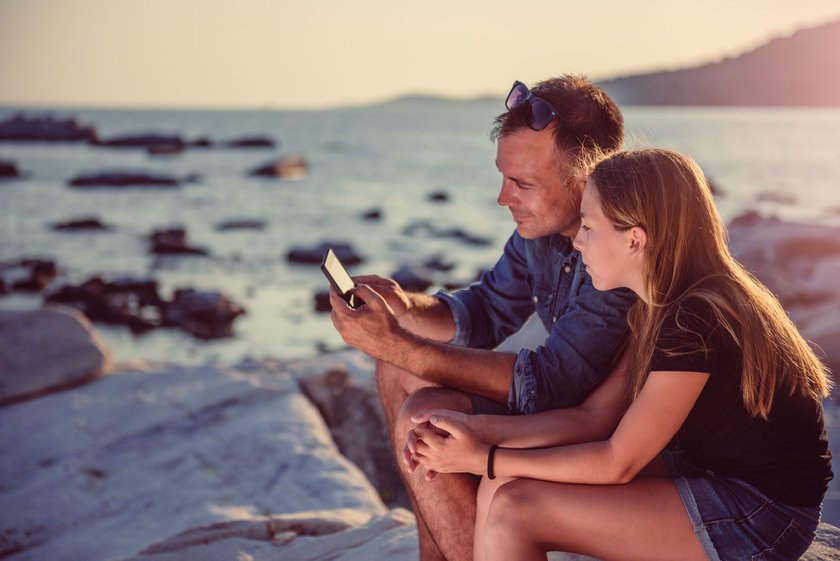 Schönen Urlaub wünschen per Whatsapp: Vater und Tochter sitzen an Steinstrand und schauen aufs Smartphone