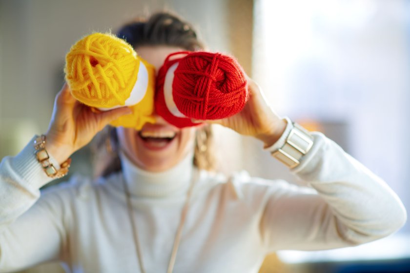 Closeup on cheerful stylish female in white sweater and skirt fool around with yellow and red knitting yarn hunks in the modern living room in sunny winter day.