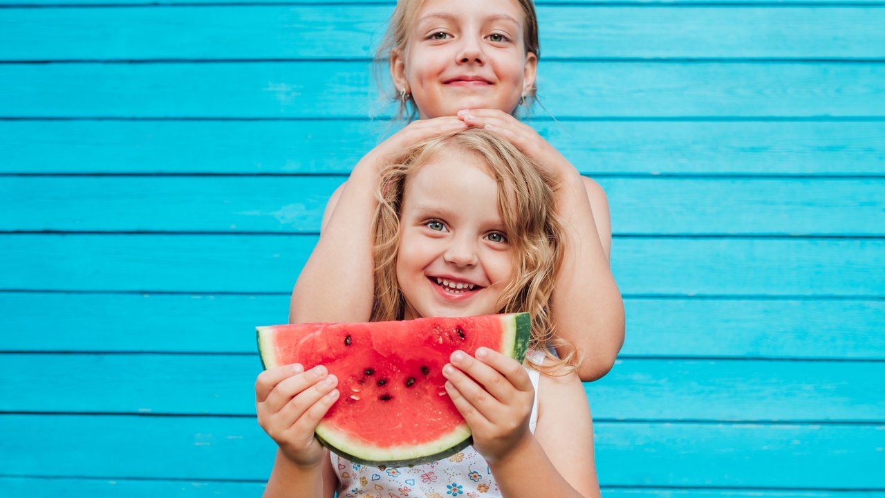 Two little sisters together with red ripe watermelon smiling. Over blue plank wall background