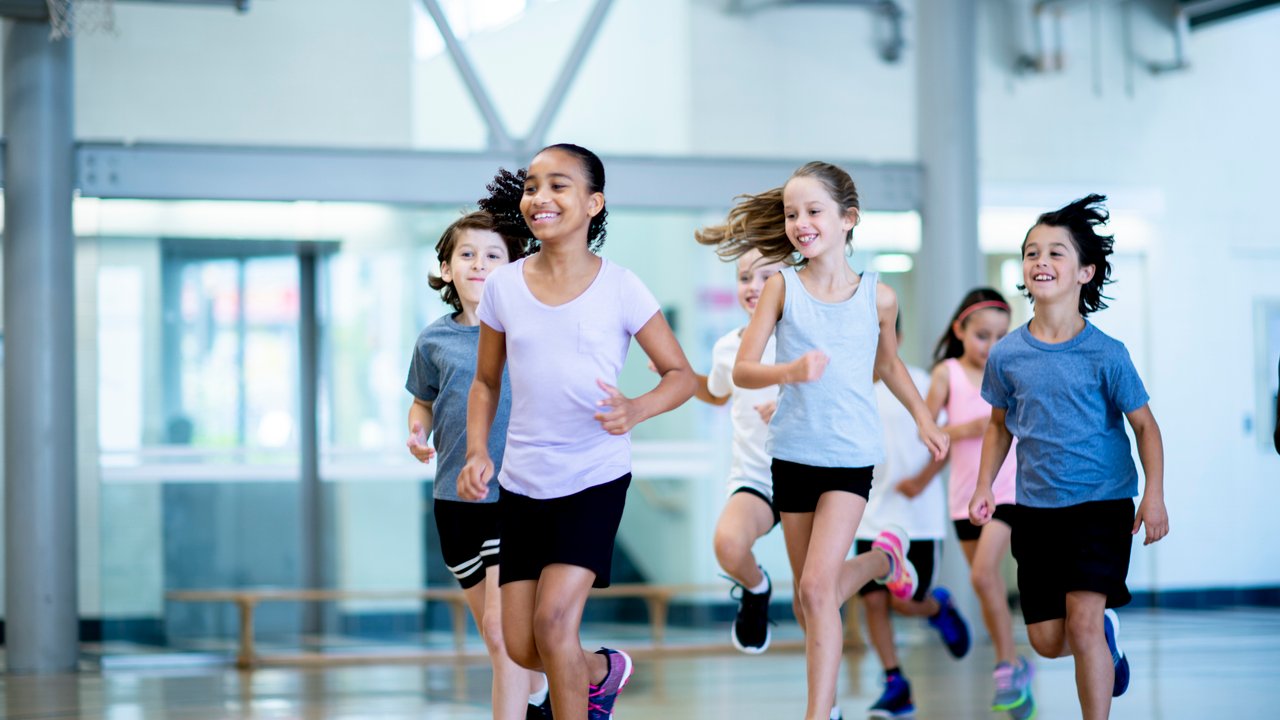 A group of diverse kids run through the gym while laughing. They are racing each other and trying to be the fastest.