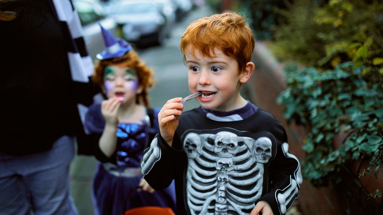 Little boy eating halloween candy while trick or treating in suburban streets with his mother and sister.