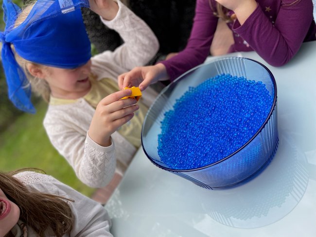 Meerjungfrauen-Geburtstag: Wasserperlen-Spiel: Kind zieht mit verbundenen Augen einen Gegenstand aus Schüssel mit blauen Wasserperlen