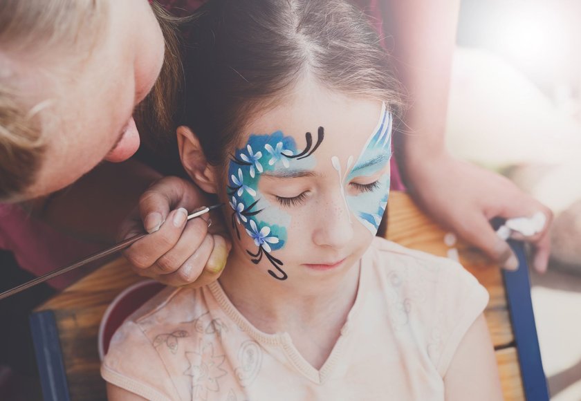 Child animator, artist's hand draws face painting to little girl. Child with funny face painting. Painter makes blue butterfly at girl's face. Children holiday, event, birthday party, entertainment.