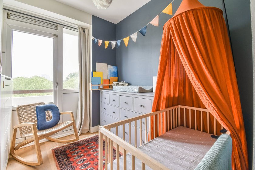 Baby crib with orange canopy placed near white drawers and armchair in light children bedroom with garland and door CasaMedia_VanNijenrodeweg858_14.jpg Copyright: xCasaxMediax