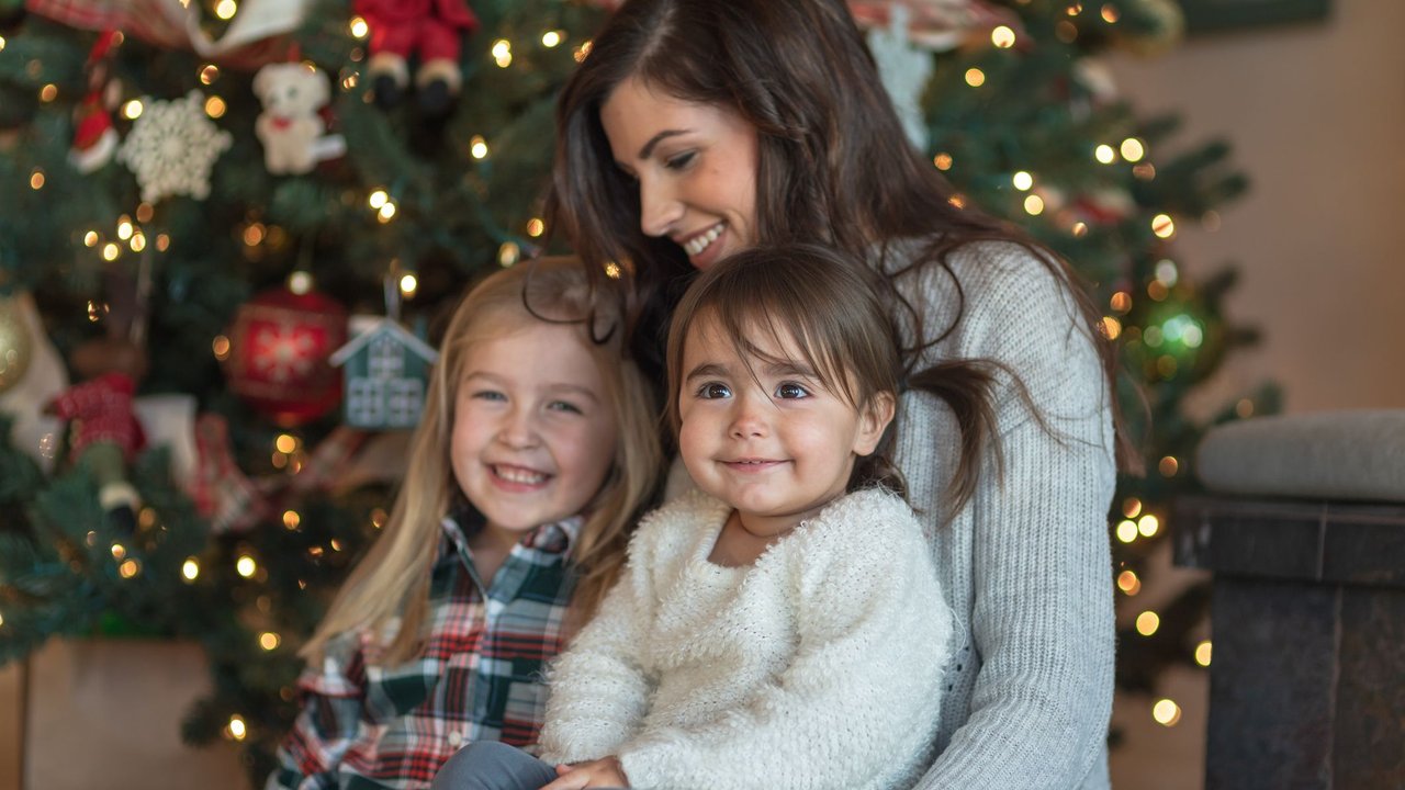 A big sister teases her little sister as they sit on Mom's lap in front of the Christmas tree in their living room. All are smiling and the two sisters are looking at the camera.