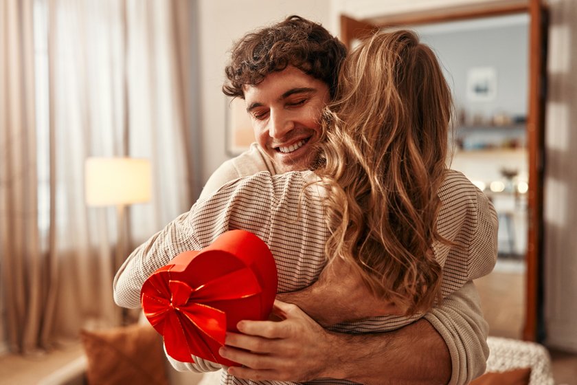 Happy Valentine's Day. A man gives a heart-shaped gift box to his beloved woman in the living room at home, the woman hugging him tenderly. Romantic evening together.