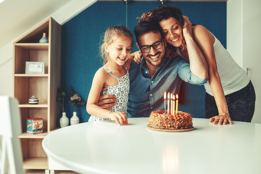 Glückwünsche zum Geburtstag Mann: Tochter, Frau und Mann umarmen sich , auf dem Tisch davor steht ein Geburtstagskuchen
