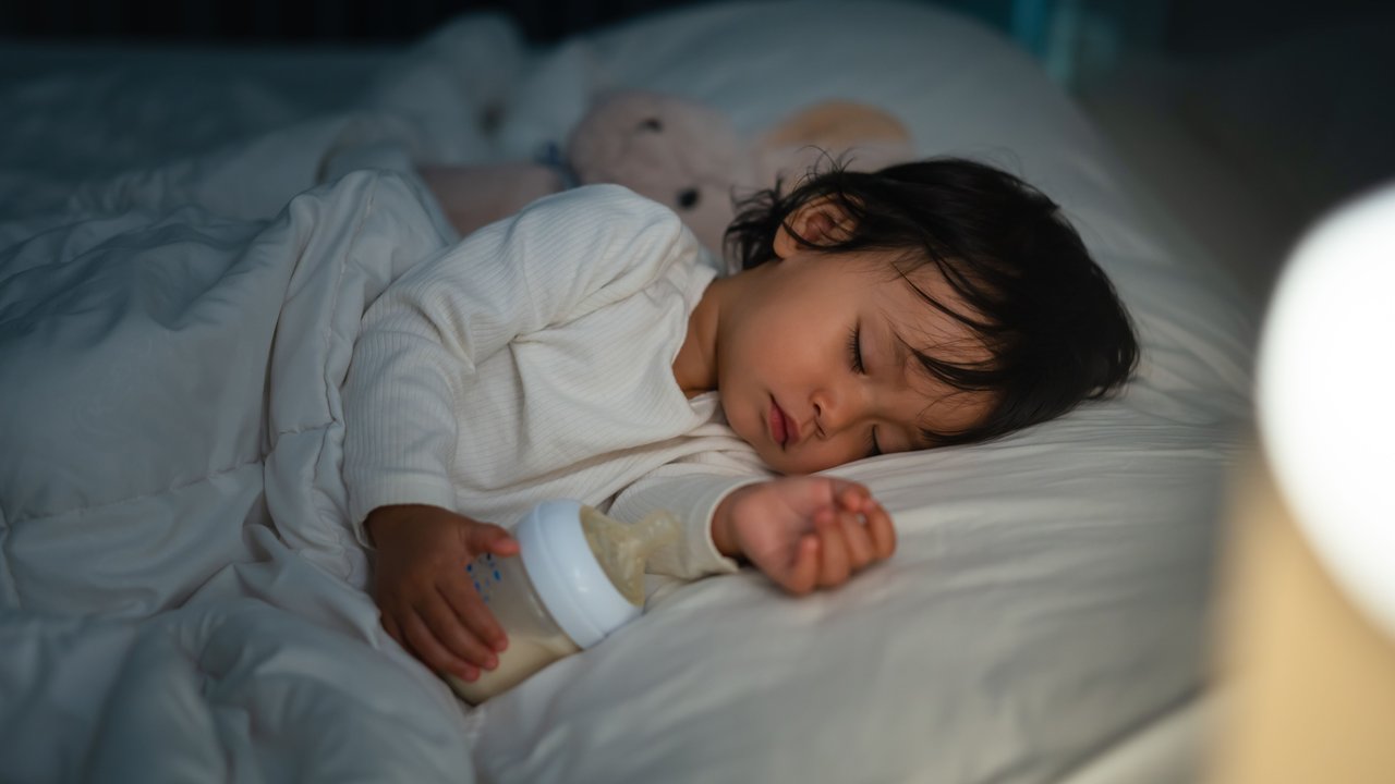 Kleinkind liegt mit Fläschchen in der Hand nachts im Bett