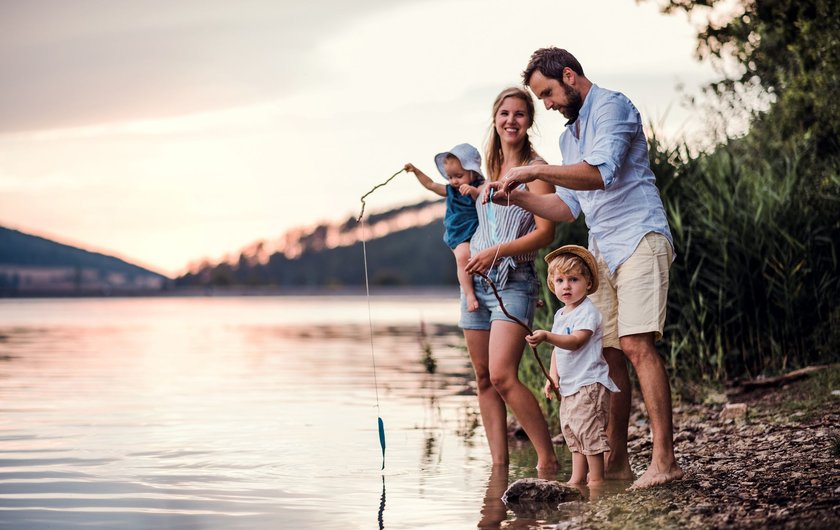 A young family with two toddler children outdoors by the river in summer, playing with paper boats.