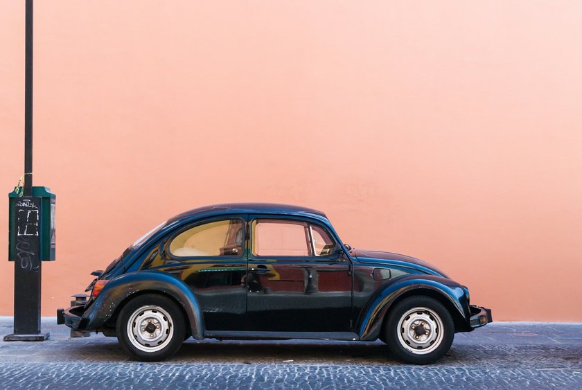 Alter, schwarzer VW Käfer vor einer rosa Wand. 