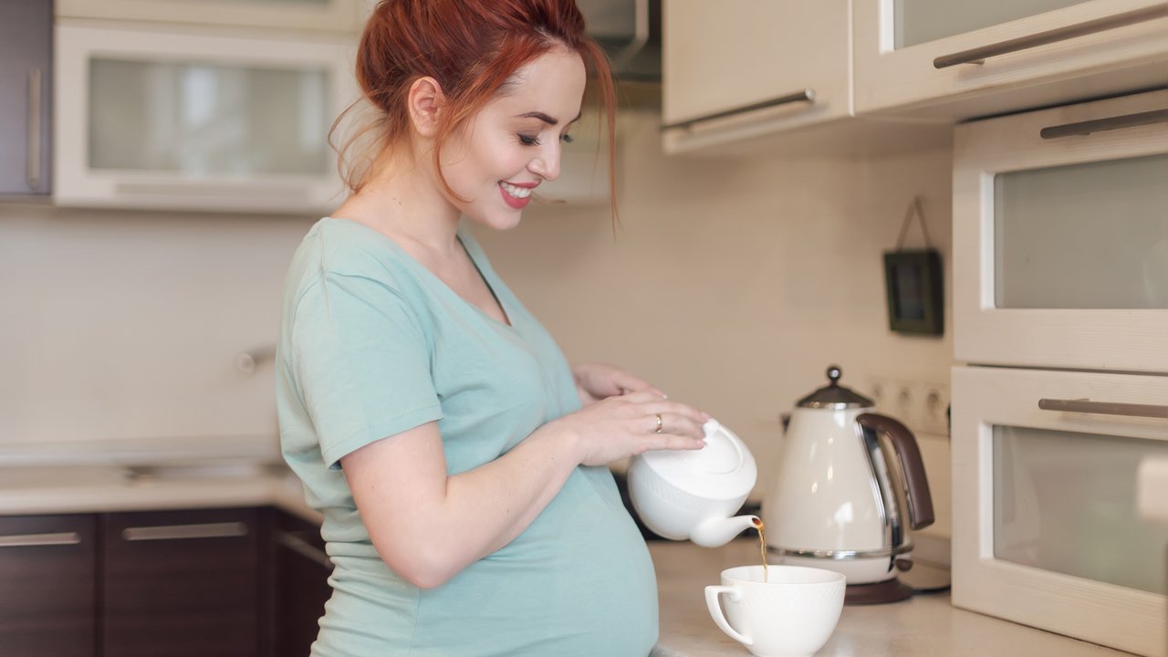Smiling pregnant woman pouring tea from a white teapot in a white cup while being in the kitchen