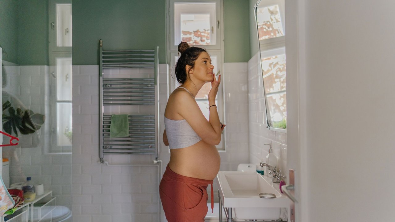 Photo of a young pregnant woman getting ready in the bathroom for the upcoming day; applying moisturizer as a part of her daily routine.