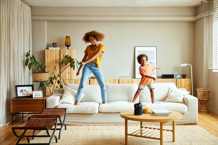 Full length of happy mother and son dancing on sofa in living room. Woman and boy enjoying at home. They are in casuals.