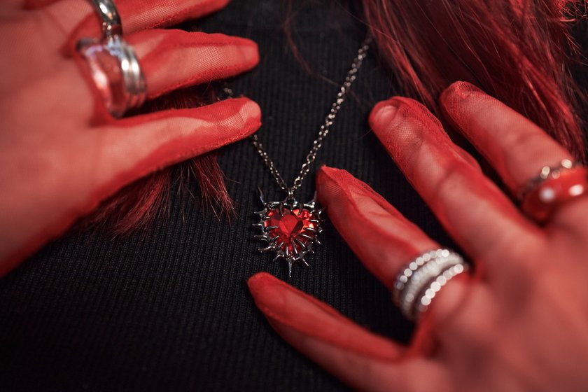 Caucasian teenage girl wearing red mesh gloves touching spiked heart pendant necklace with both hands, showing multiple rings on fingers, creative Gen Z fashion style at home