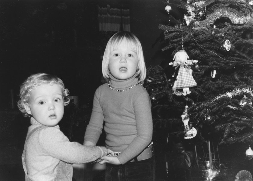 Ein Foto aus den 70er Jahfren zeigt einen Bruder und eine Schwester, die sich an den Händen halten und nebeneinander neben einem Weihnachtsbaum stehen