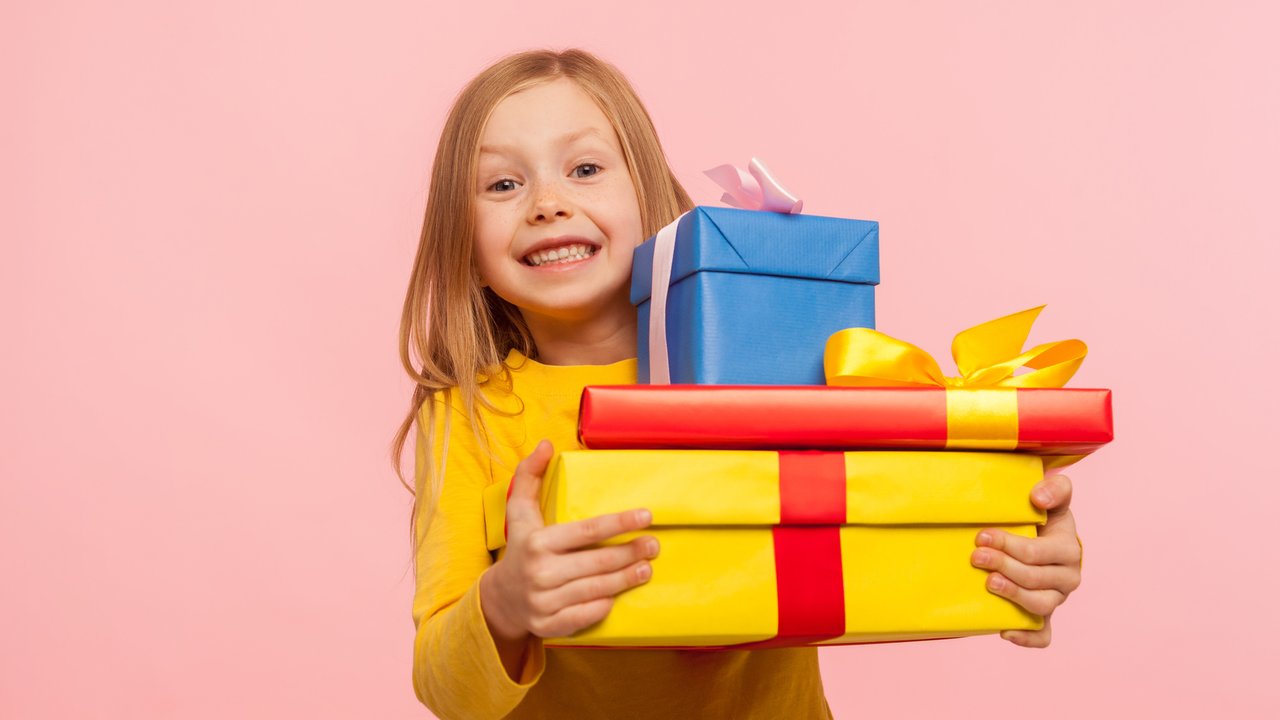 Delighted little girl embracing lot of gift boxes and smiling at camera with expression of sincere childish happiness, enjoying perfect birthday with many presents. indoor studio shot, pink background