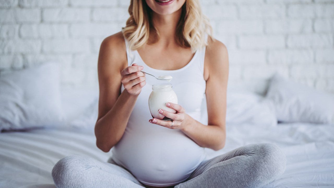 Cropped image of attractive pregnant woman is sitting in bed and eating yogurt. Healthy food concept. Last months of pregnancy.