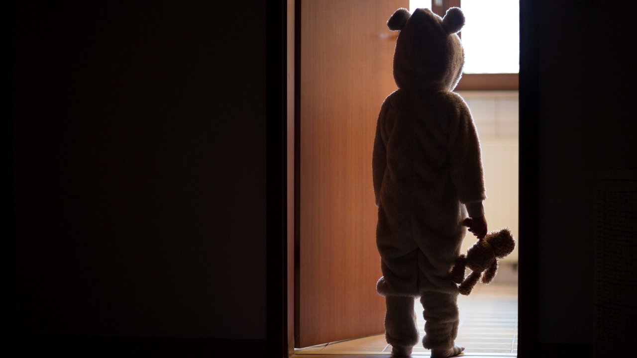 Child in bear costume, standing in a darkened doorway, looking through to the light