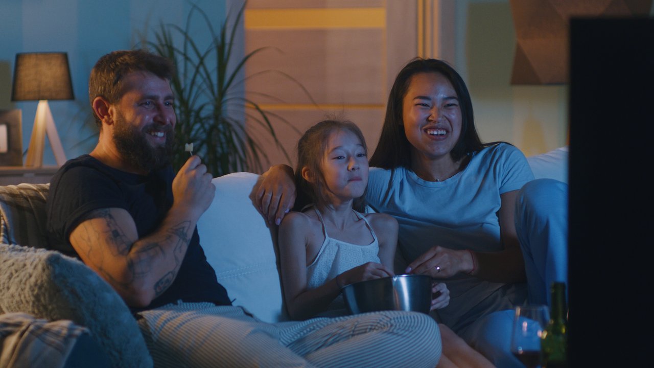 Happy diverse parents and daughter laughing and eating popcorn while relaxing on couch and watching comedy film in evening at home together