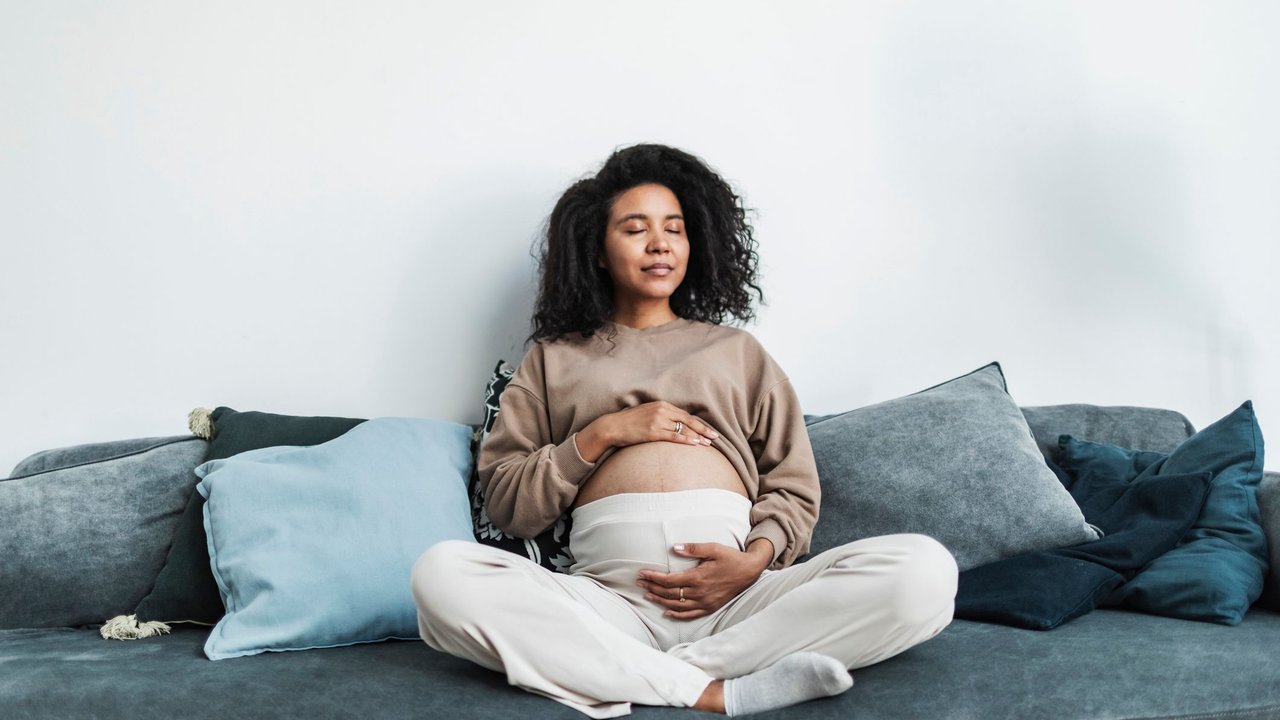 Pregnant black woman sitting on sofa with closed eyes, touching her belly and smile