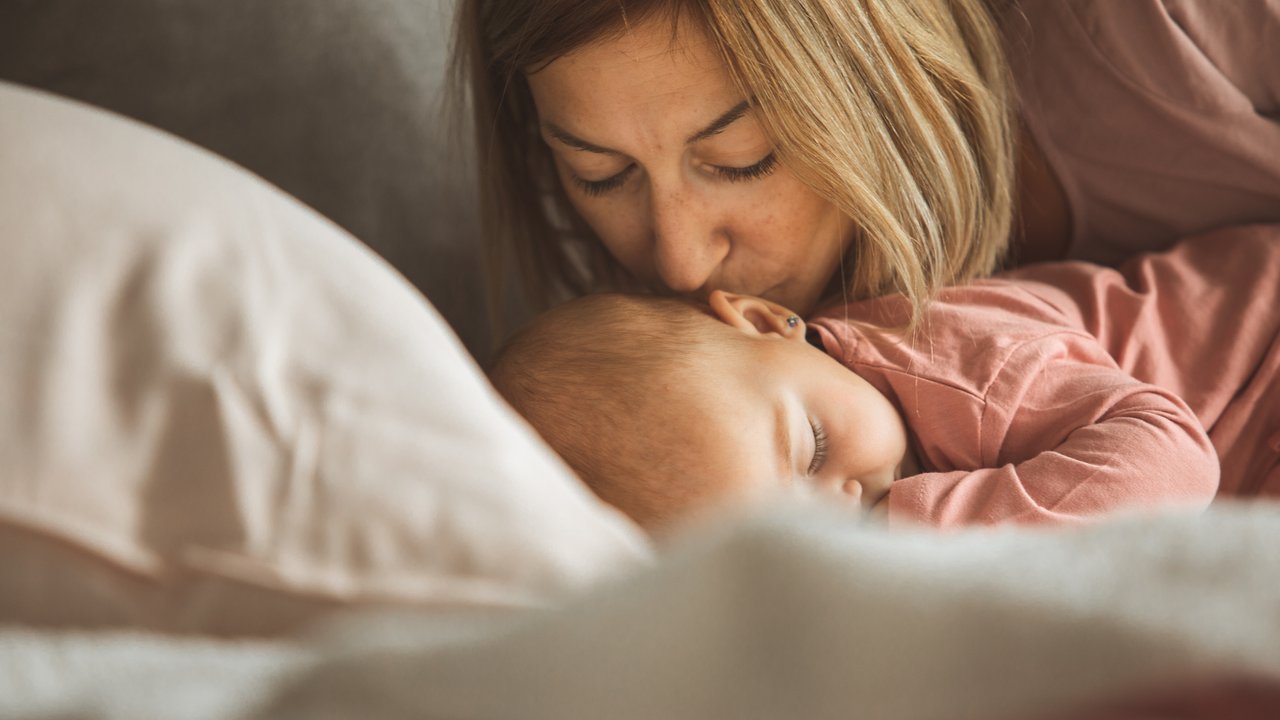 Close-up of a mother kissing her baby in the head while she is asleep on the bed.