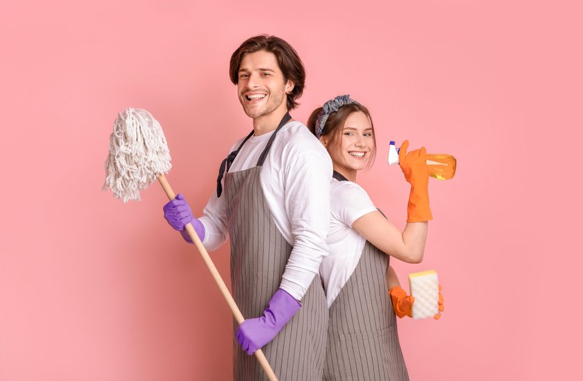 Professional Cleaning Service. Portrait Of Smiling Man And Woman With Household Tools In Hands Standing Back To Back Over Pink Studio Background