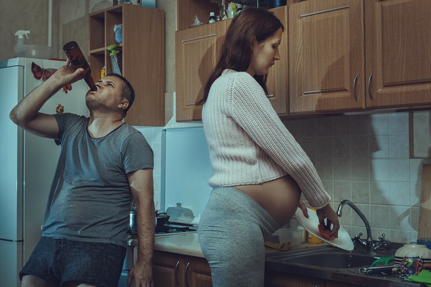 Absurdes Frauenbild hochschwangere Frau wäscht ab während der Mann ein Bier trinkt