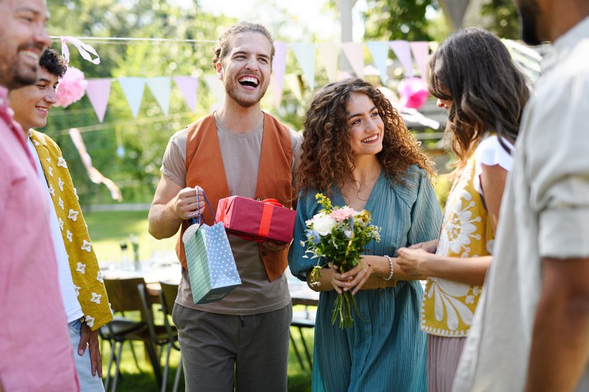 Was schenkt man zum Polterabend: Mann hält Geschenke, Frau einen Blumenstrauß in der Hand bei Gartenparty mit Freunden
